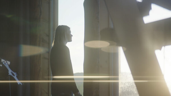 Micah Atkins stands at a window looking out as the sun begins to set. There is a glare from the sun that cuts across the screen.